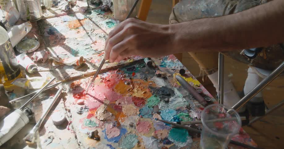 Painter sitting in front of a canvas panel on an easel fills a brush with paint on a pallet