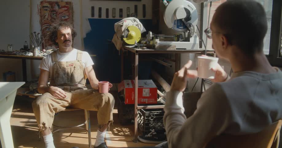 Two men sit in artist studio with coffee mugs conversing in sign language