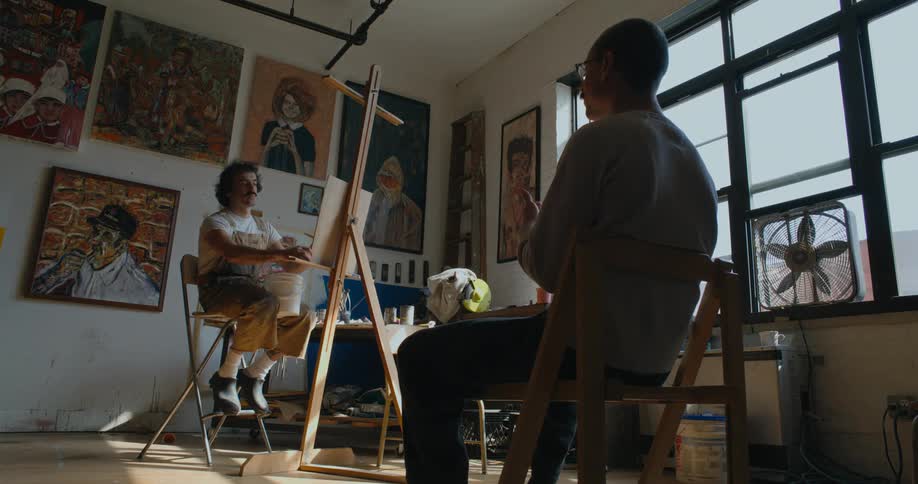 Artist paints sitting in front of easel in studio while model uses sign language