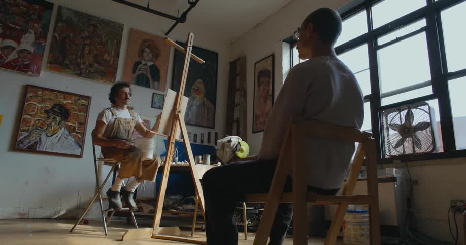 An artist talks with a his model while painting his portrait indoors during the day