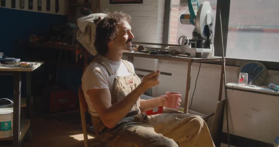 Man with mustache wearing overalls sitting in a studio talking with sign language