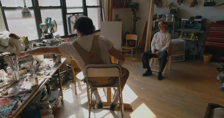 A man is sitting in a chair next to a table covered with paint brushes and art supplies as he talks to another man sitting in a chair across the room