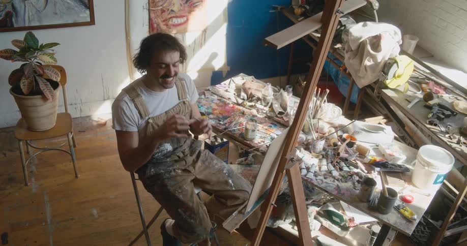 A painter sits in front of a painting and talks as he moves his hands and paint brush in the air and then paints
