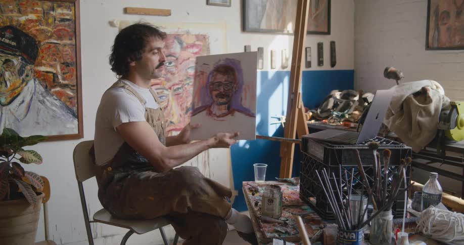Painter sitting at easel picks up small painting and discusses it in sign language