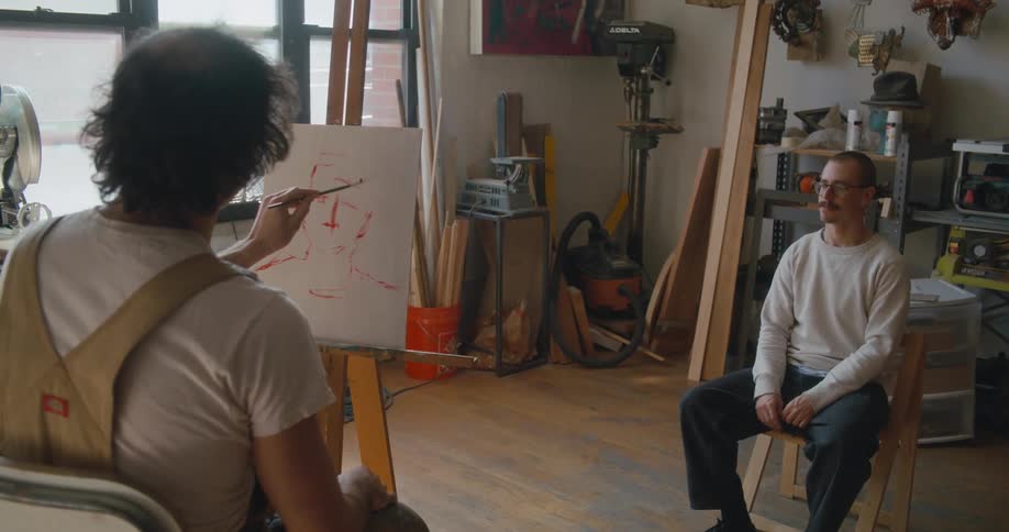 Male painter paints portrait of man sitting on chair in studio