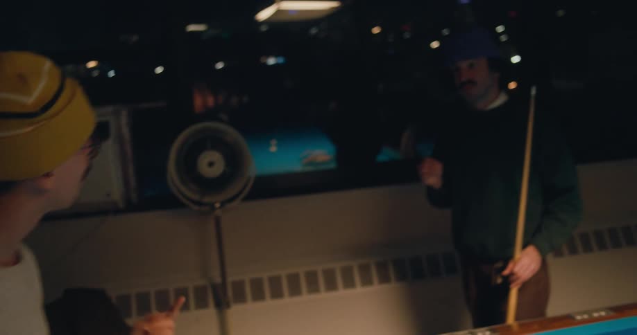 A pair of men talk with one another while one of them uses a pool rack to place the billiard balls in the center of the pool table