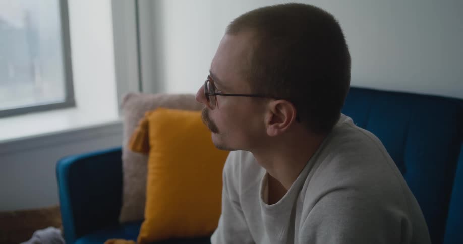 Man with a mustache wearing glasses sitting on a couch in the living room at home