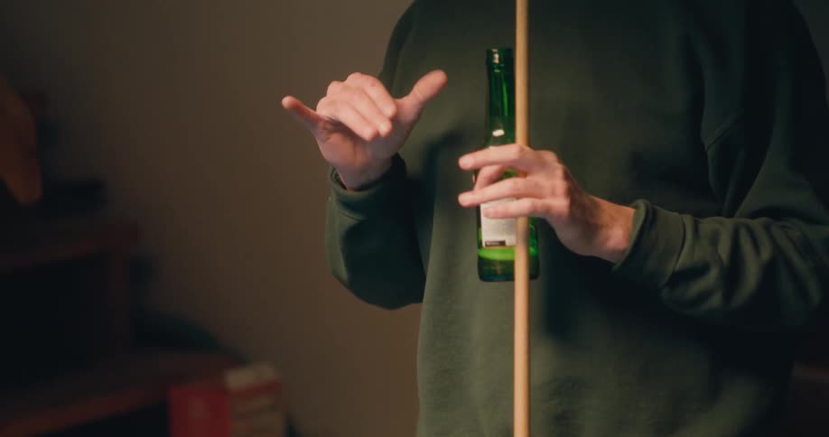 A man is holding a pool cue stick and a beer bottle and he makes a hand motion and drinks his beer