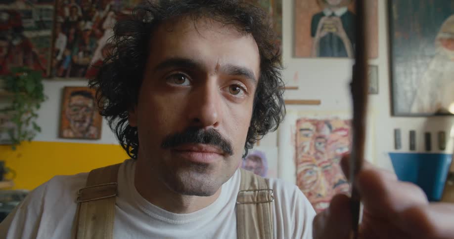 Artist stands against wall full of artwork contemplating his work in progress before painting again