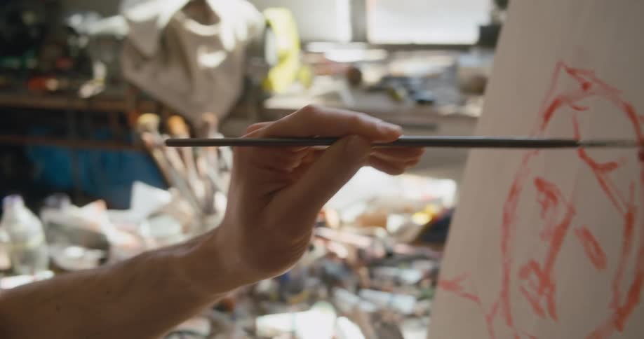 A man is sitting down painting a picture on an easel and he has a table full of brushes and paint next to him