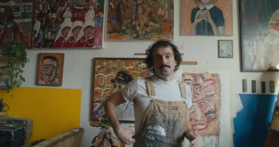Male artist finishes painting then stands and moves to pose against wall with finished paintings