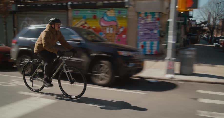A man wearing a bike helmet is riding a bike through town passing cars and buildings