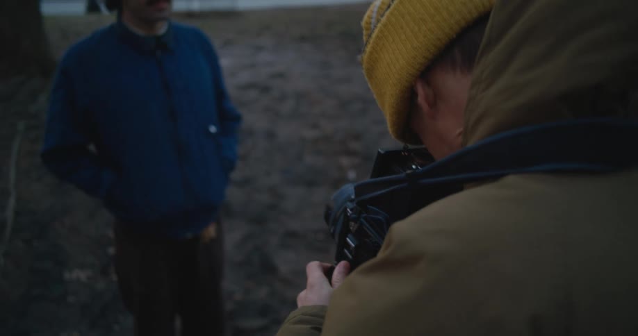 A man is holding a camera and taking a picture of another man standing near trees in a park