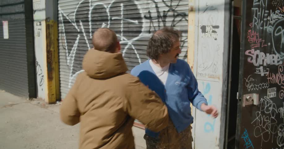 A man uses his phone outside a door until another man exits and greets him before leading him through the door