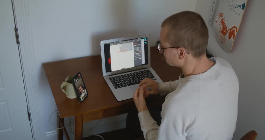 Man sits at desk with computer while signing with another man via video call on a smartphone propped against a coffee cup
