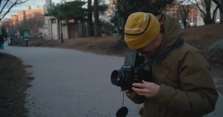 Man with yellow wooly hat glasses mustache and brown coat takes photo in public park