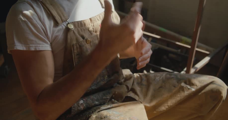 Man wearing paint splattered overalls sits in chair and uses sign language