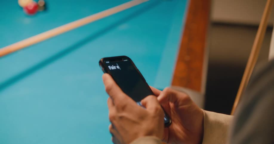 A man types on cell phone and then hands the phone to another man who also types as they play pool