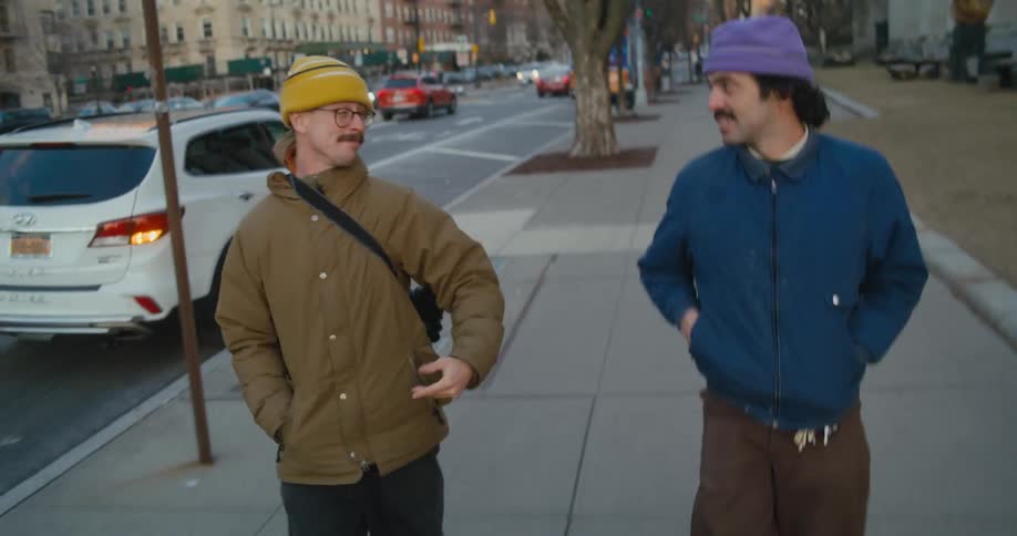 Two men wearing jackets and hats walk on the city sidewalk as one of the men smiles and points to the street on his right