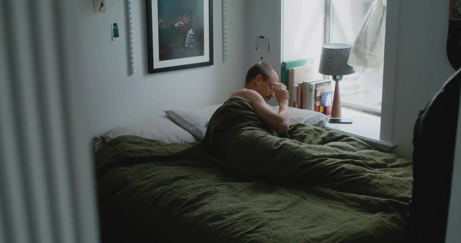 A man is lying in bed under the covers and he sits up and puts on his glasses and looks at his phone