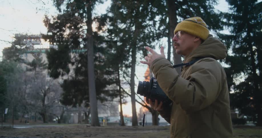 A man is standing outside near trees in a park holding a camera and taking a picture as he holds up his fingers