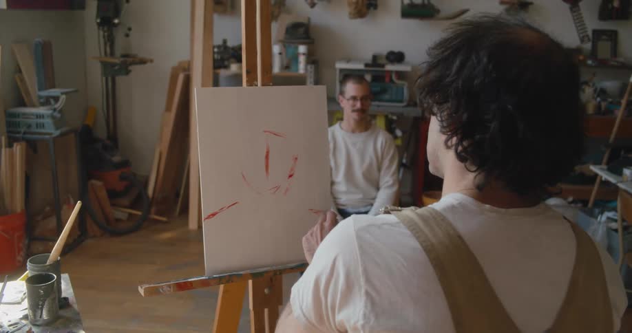 A man sitting in a studio workshop while an artist begins to paint on an easel