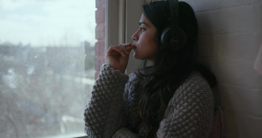 A woman listens to headphones while sitting next to a window