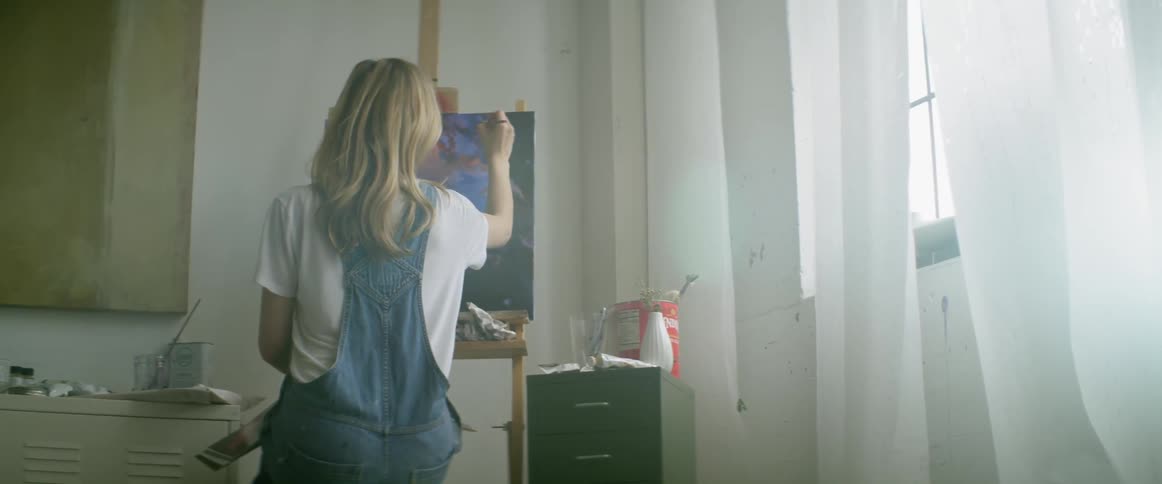 A woman in blue overalls is painting indoors