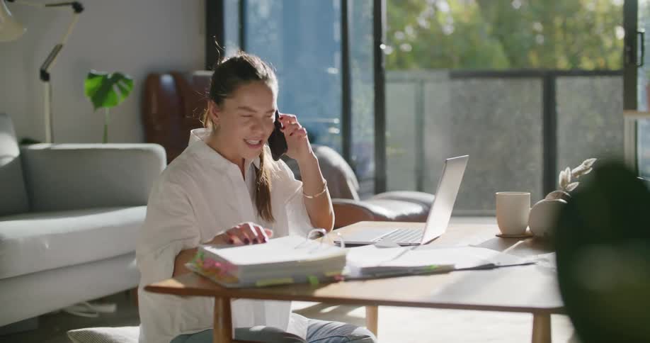 A woman sits on the floor talking on the phone as she flips through a large binder and uses her laptop
