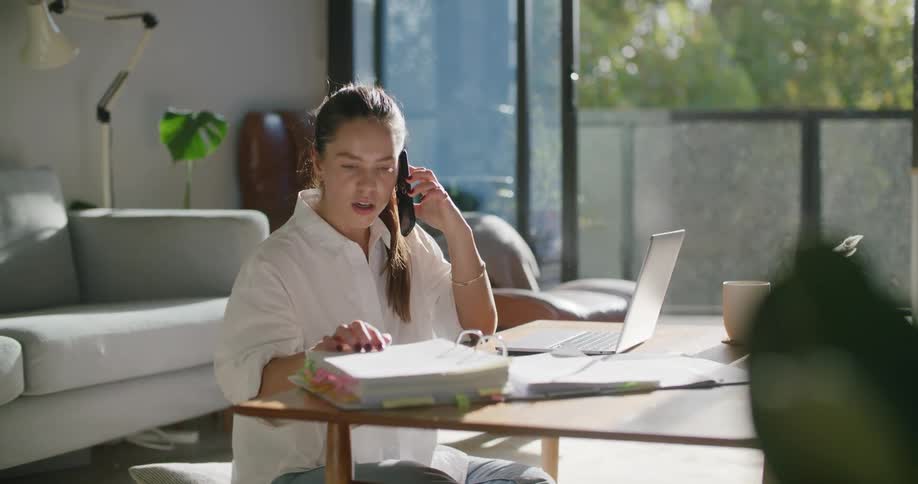 A woman on a smart phone sitting at a table typing on a laptop with a couch in the background