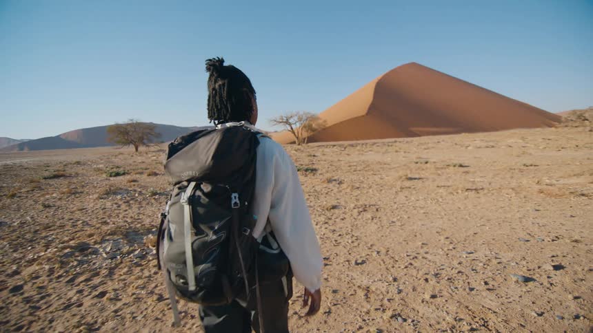 A woman hikes through the desert towards a large sand dune in the distance