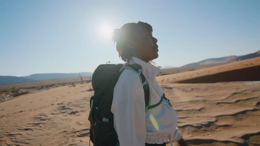 A woman wearing a pack walks on the sand with the sun shining down