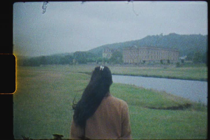A woman in a long coat walks through a field next to a pond on a cloudy day