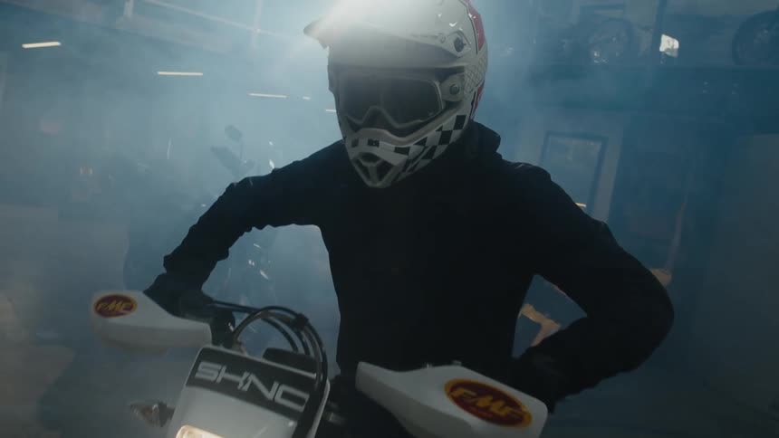 A light flashes on a motorcycle and rider wearing a helmet sitting on the motorcycle in a smoke filled room