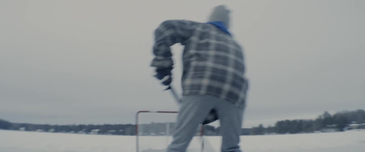A teenager skates and shoots a puck into a net on a frozen lake while others stand in a group talking