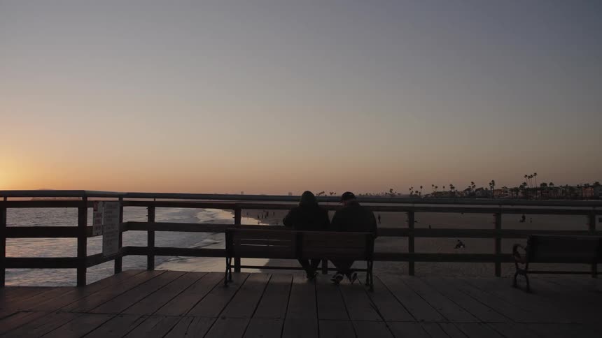Elderly man and woman sit on a bench and look out at the ocean and beach at dusk