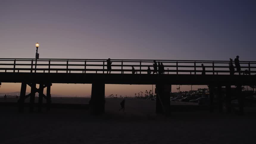 The silhouettes of multiple people are seen as they walk along a pier in low daylight