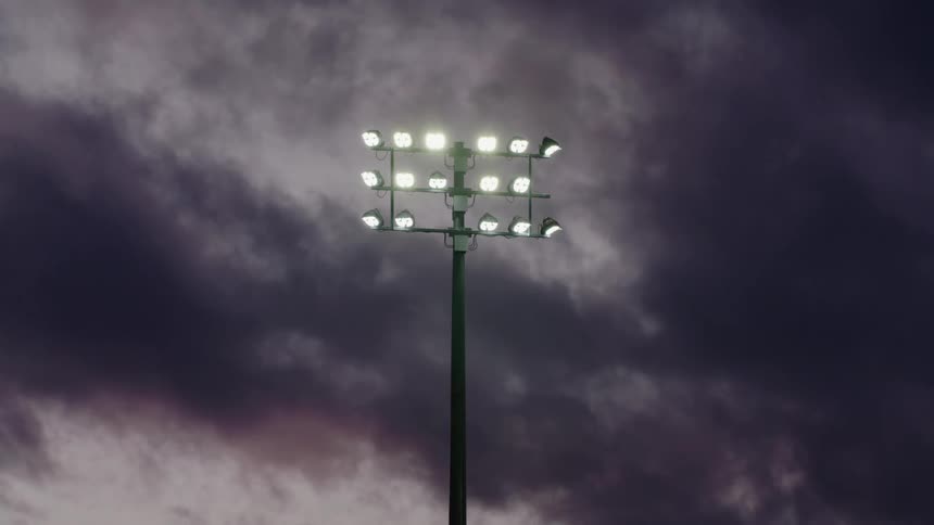 An array of stadium light shine brightly beneath a cloudy sky