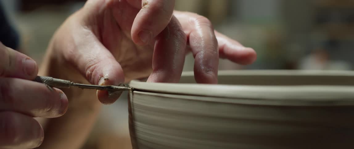 A man holds a blade to create lines on pottery on a spinning wheel