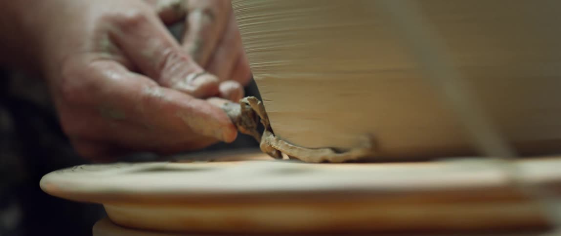 Close up of a mans hands uses a scraper to mold pottery over a wheel
