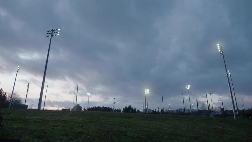 Sports field lights shine under cloudy sky as red tints horizon