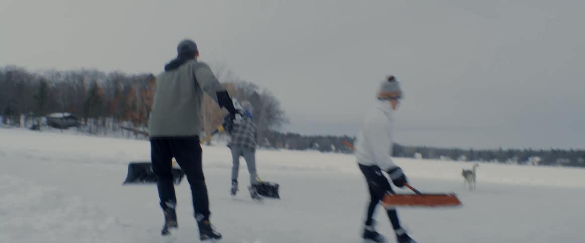 A group of men and women use brooms to clear snow off a iced over lake