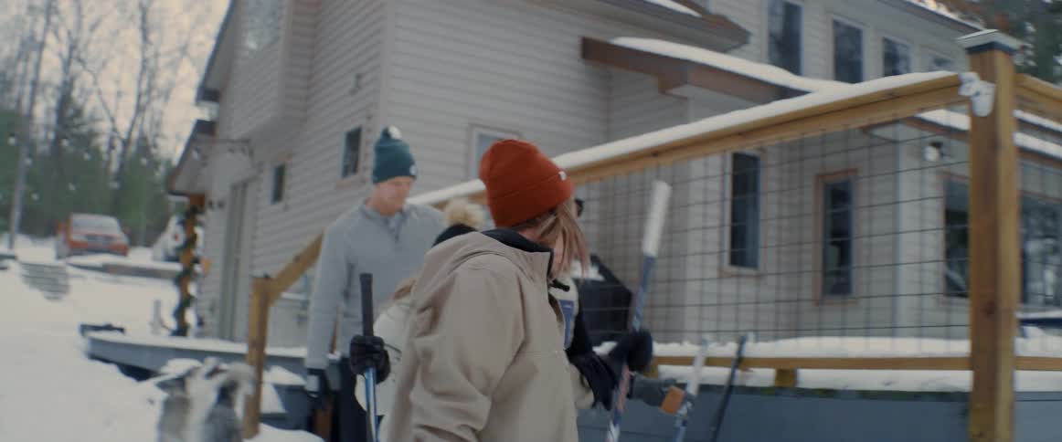 Happy family of four wearing winter clothes outside the house during a snowy day grabbing the hockey sticks that are leaning against the fence
