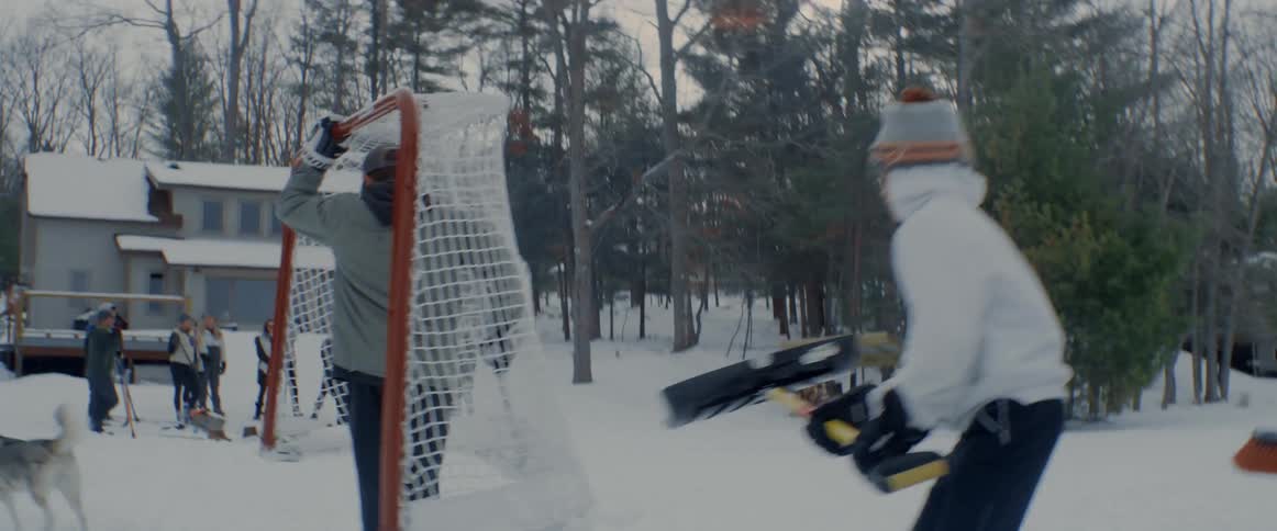 Teenaged boy carries a hockey net as he skates with others carrying shovels and a broom heading towards a group of people waiting at an outdoor rink
