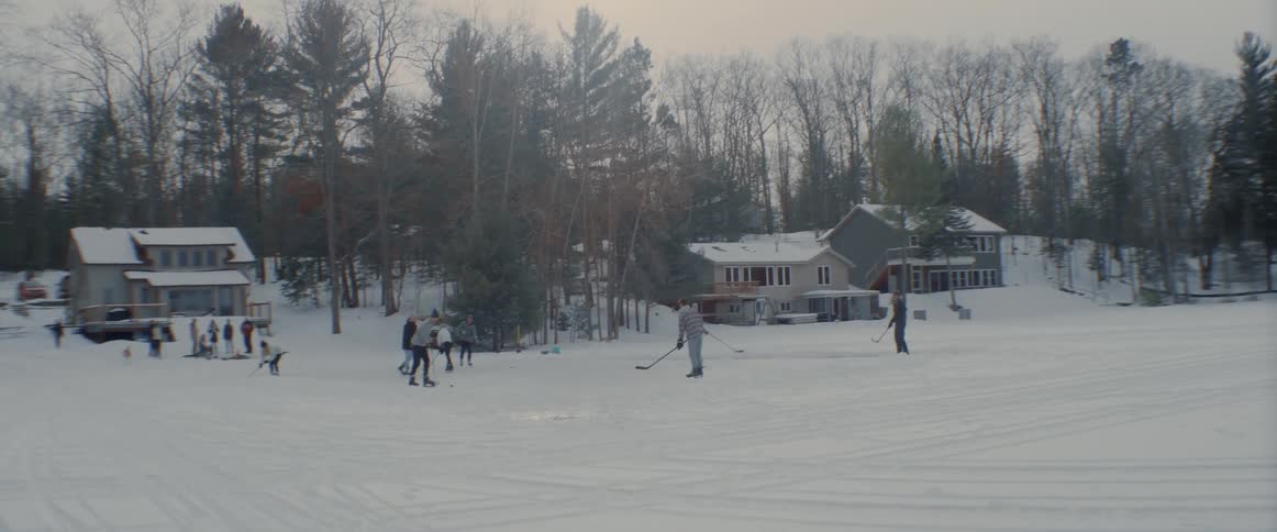 A group of people are outside playing hockey in a neighborhood field on a winter day and others are watching