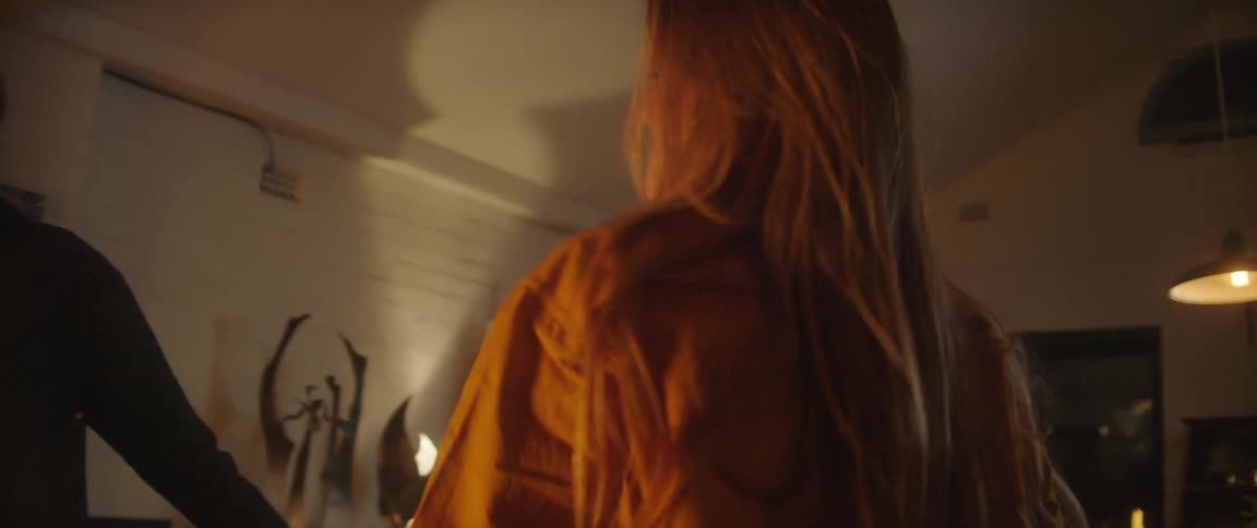 Blonde woman with brown jacket and black top dances with man wearing denim jacket in living room at night