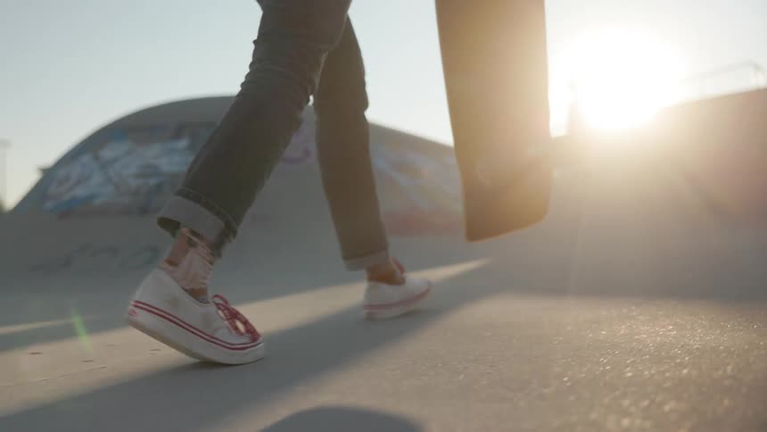 Young woman wearing sneakers carries a skateboard over a skate park at sunset