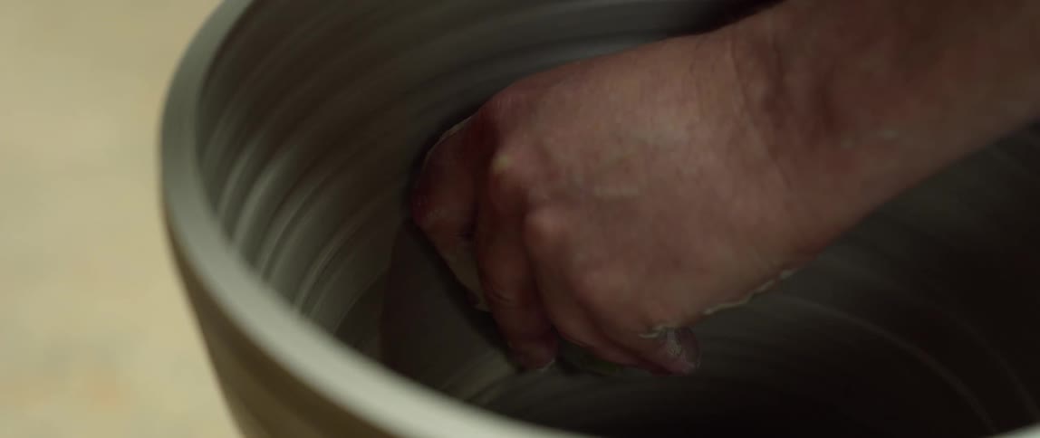 Close up of a man shaping up a clay vase over a pottery wheel with a scraper