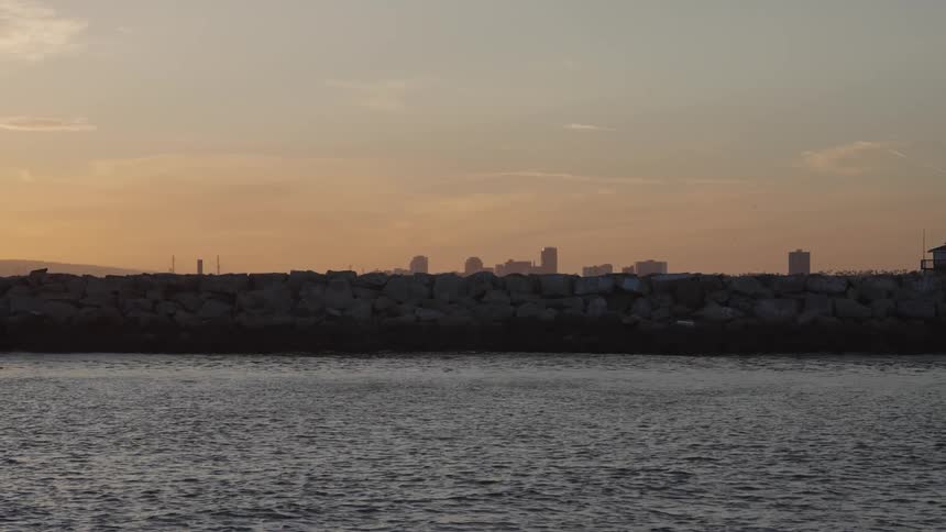 A jetty projecting into water on the coast in front of the city skyline at sunset
