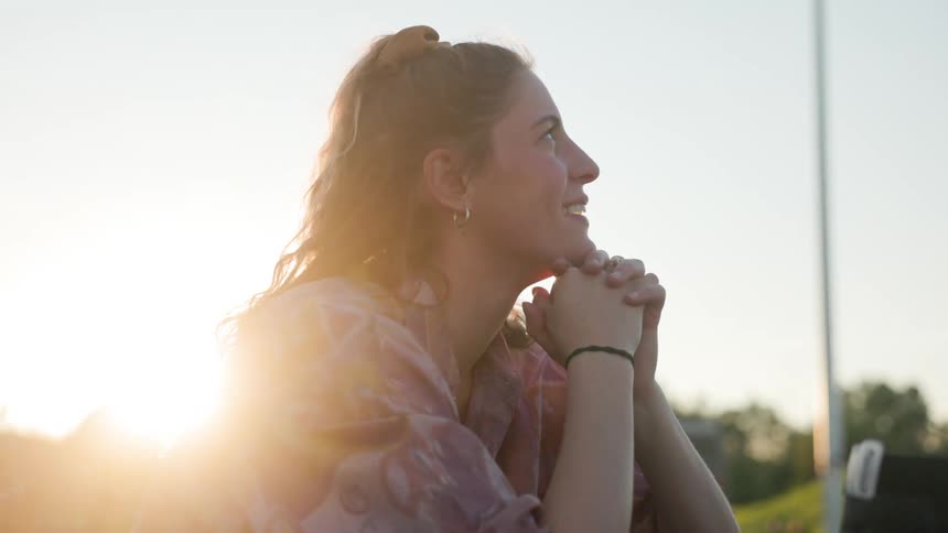Happy white woman outdoors during a sunny day holding her hands together next to her face looking around and smiling
